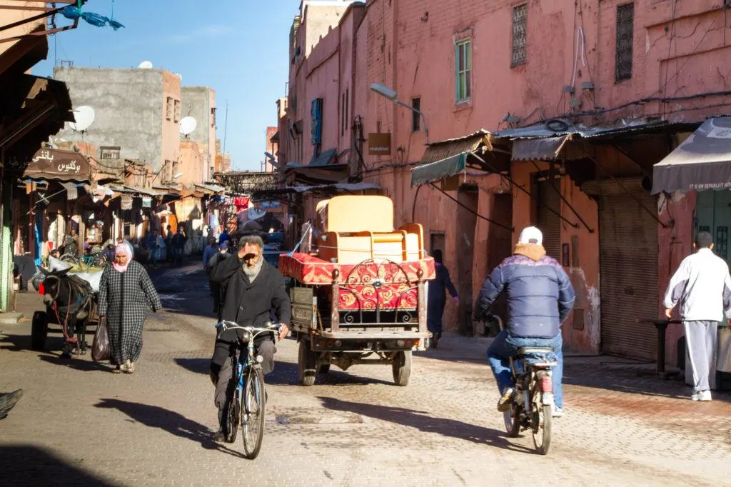A busy Marrakech street scene with donkeys pulling carts, men on bikes, and a women carrying a shopping bag.