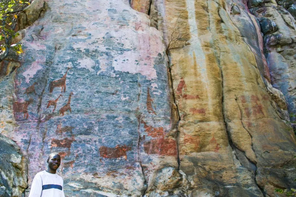 San guide standing below a huge rock with ancient animal figures, including giraffes and antelope at Tsodilo Hills, Botswana.